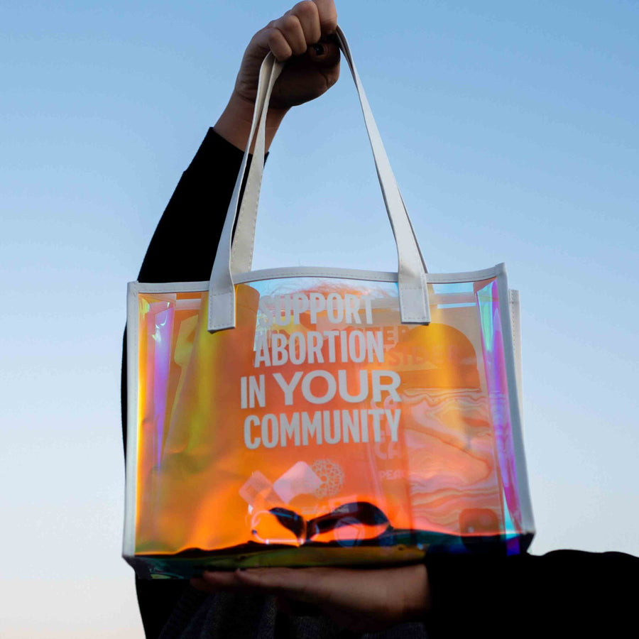 Two hands hold an iridescent, translucent, vinyl tote bag against to the sky during golden hour, a warm tone reflecting on the bag's surface. White imprint reads SUPPORT ABORTION IN YOUR COMMUNITY with the NNAF logo below.