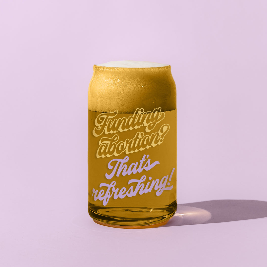 An amber-colored glass with yellow and purple script that reads "Funding abortion? That's refreshing!" Glass is filled with a foamy beverage, like kombucha or beer, and sits on a lavender background.