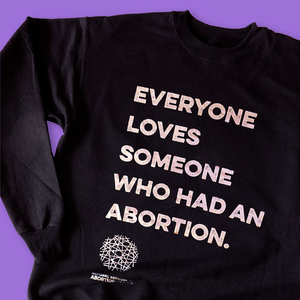 Photo of a black crewneck sweatshirt on a purple background. The shirt is imprinted with the phrase, "Everyone loves someone who had an abortion," and the National Network of Abortion Funds logo, in shiny silver foil.