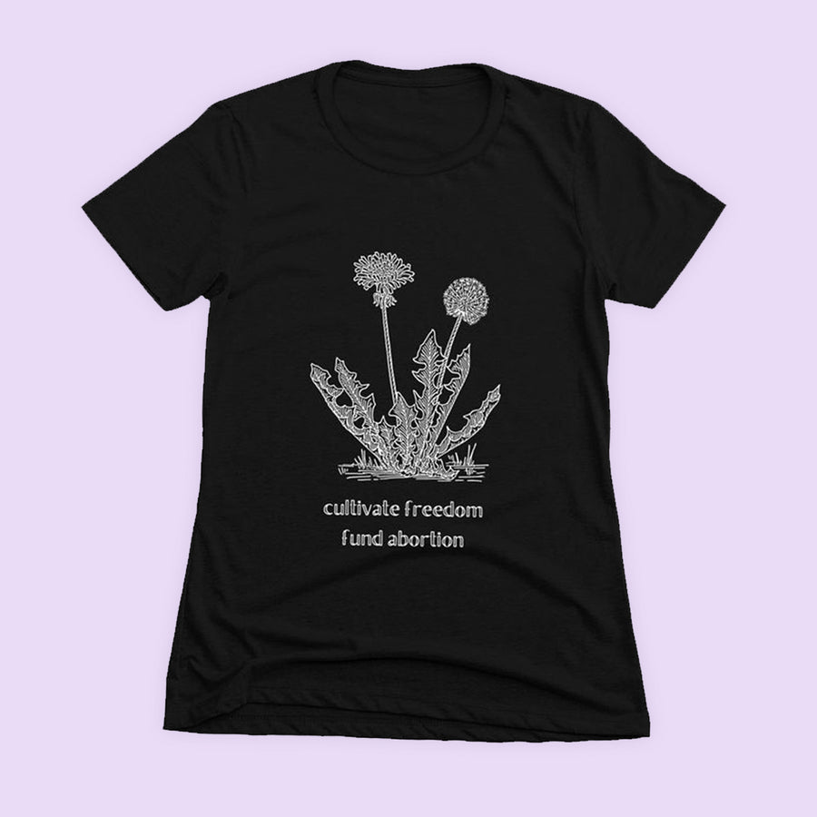 Black short sleeved tee on a lavender background, the tee's white print reads "cultivate freedom, fund abortion" and features an illustration of a dandelion in bloom.