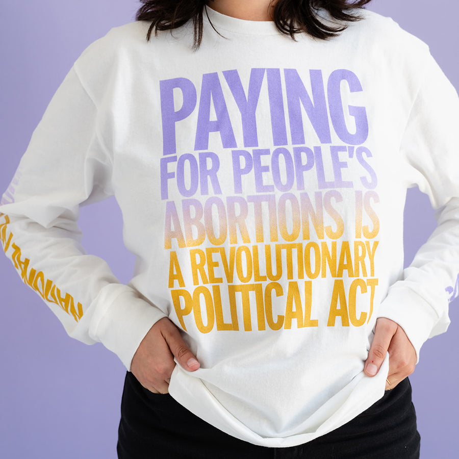 A person with wavy brown hair sports a long sleeved white tee that reads PAYING FOR PEOPLE'S ABORTIONS IS A REVOLUTIONARY POLITICAL ACT, with their hands on their hips. The model's face is cropped out of frame.