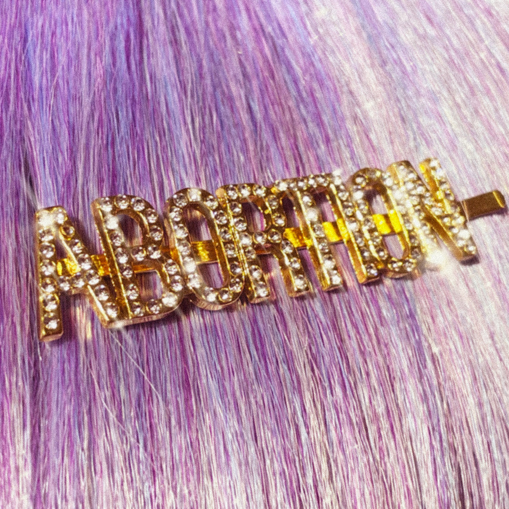 A gold hairpin with sparkly rhinestone letters that read ABORTION. The pin is displayed in straight hair that is lavender in color.