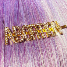 Load image into Gallery viewer, A gold hairpin with sparkly rhinestone letters that read ABORTION. The pin is displayed in straight hair that is lavender in color.
