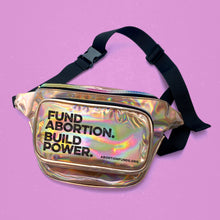 Load image into Gallery viewer, Photo of a gold holographic vinyl fanny pack with a black nylon strap, imprinted with the text, &quot;Fund Abortion. Build Power,&quot; on a light fuchsia background.
