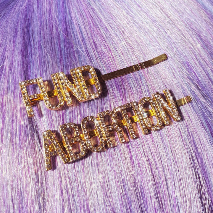 Two gold hairpins with sparkly rhinestone letters that, together, read FUND ABORTION. The pins are displayed in straight hair that is lavender in color.