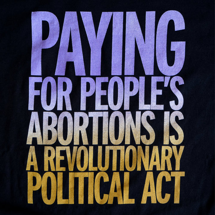 Black jersey fabric imprinted with text that reads "Paying for people's abortions is a revolutionary political act" in a purple to gold gradient.